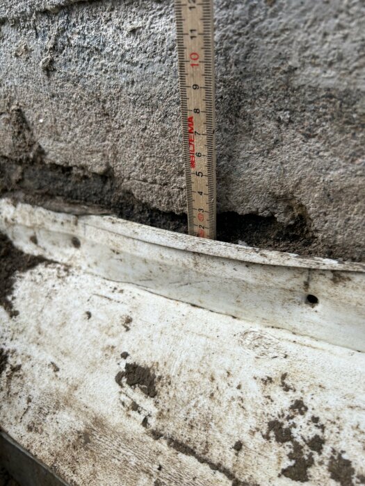 Mätstock placerad mot en mögelskadad vägg med synlig betong, isolering och byggplast vid en dräneringsgrävning.