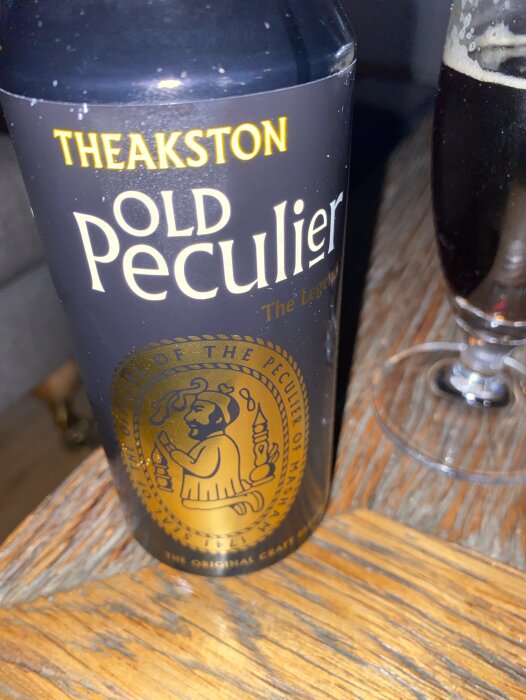 Theakston Old Peculier ölflaska bredvid ett delvis fullt ölglas på ett träbord.
