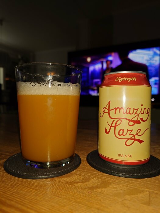 Ölglas fyllt med skummande öl och en burk IPA, Amazing Haze, på ett bord med suddig TV i bakgrunden.