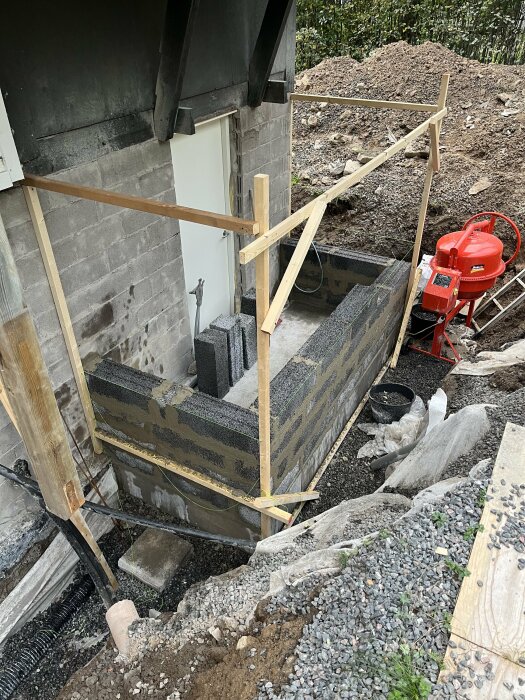 Byggprojekt med gjutform och armeringsnät, betongblandare och makadamgrund utförs vid husvägg.