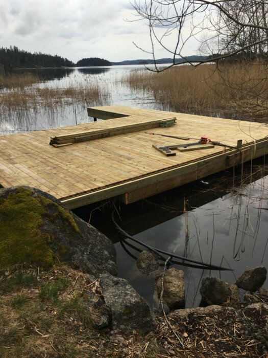 Nybyggd träbrygga vid sjö, synliga kablar och reglar mot berg, verktyg på bryggan.