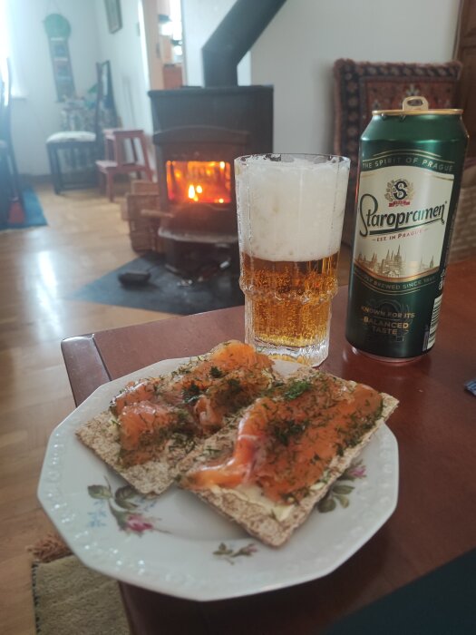 Mellanmål bestående av gravlax på knäckebröd och ett glas öl framför en öppen spis.