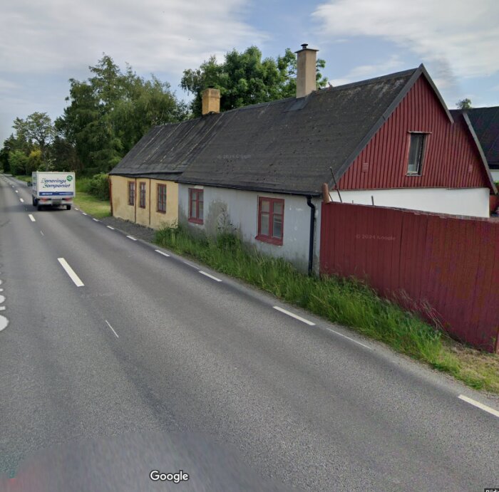 Gammalt hus med röda och vita väggar nära en trafikerad väg, omgivet av grönska.