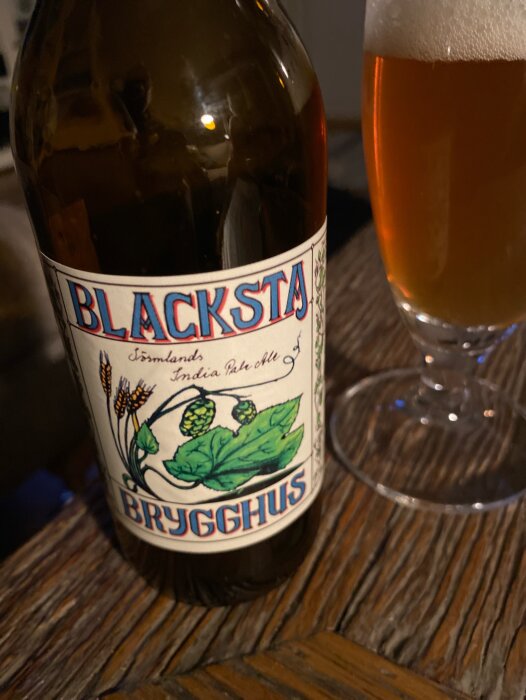 Flaska av Blacksta öl bredvid ett glas fyllt med ljust skummande öl på ett träbord.