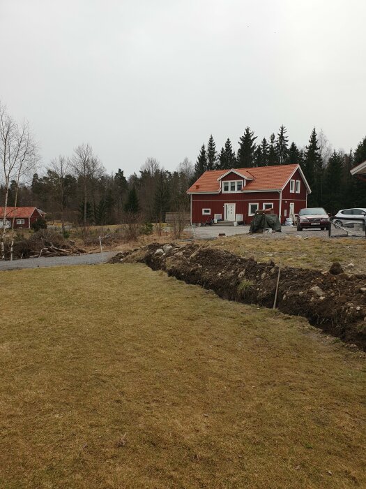 Nygrävt dike i kant av gräsmatta nära röd hus med dräneringsarbete pågår.