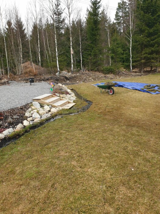 Synlig gräsmatta med dränageproblem, en upphöjd köksträdgård halvmåneformad med stenar, en skottkärra och blå presenning.