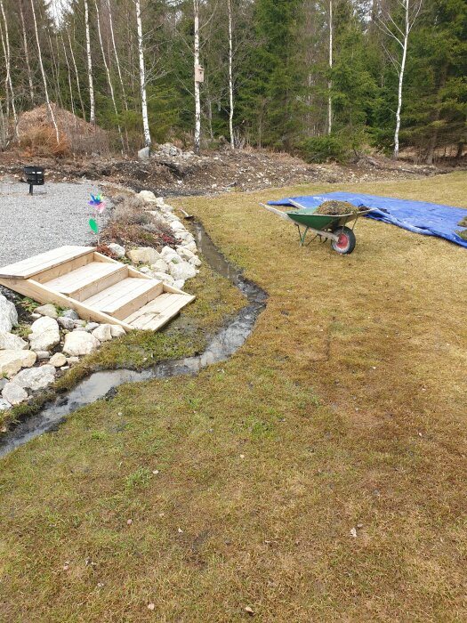 Genomblöt gräsmatta med grävd ditch, vattenproblem vid upphöjd köksträdgård och skottkärra.