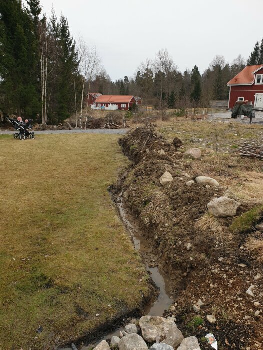 Nygrävt dike i trädgård med lera och stenar för att förbättra dränering nära en gräsmatta.