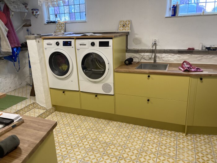 Tvättstuga med Epoq-skåp i grönt, tvättmaskin, torktumlare och mönstrat golv.