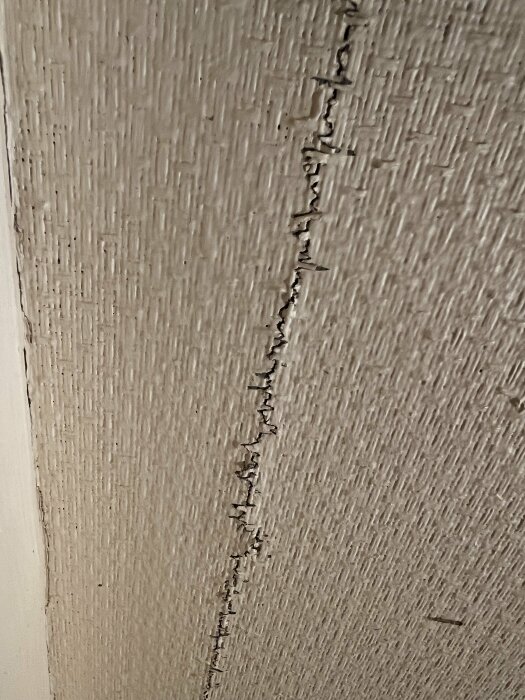 Spricka i övermålad vävtapet på vägg med synlig reparation och trälist i nedre delen.