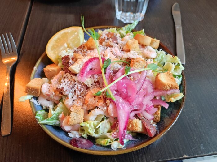 Caesarsallad med räkor, krutonger, parmesan, picklad lök och citron på en mörk tallrik.