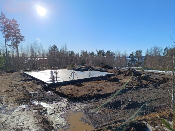 Nybyggd husgrund med rörinstallationer på en byggarbetsplats, omgiven av lerig mark och skog.