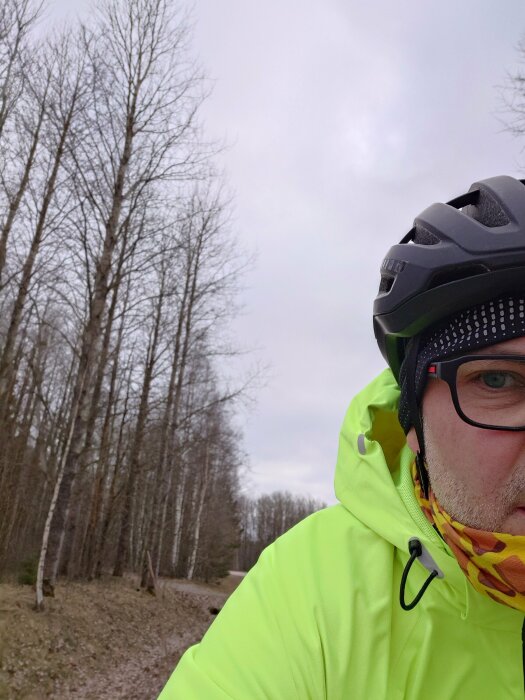 Person i cykelhjälm och neongul jacka, halvt ansikte synligt, med skog och stig i bakgrunden.