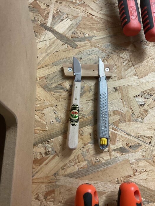 En täljkniv och en märkkniv på ett spånskivegolv med spån och verktygshandtag i förgrunden.