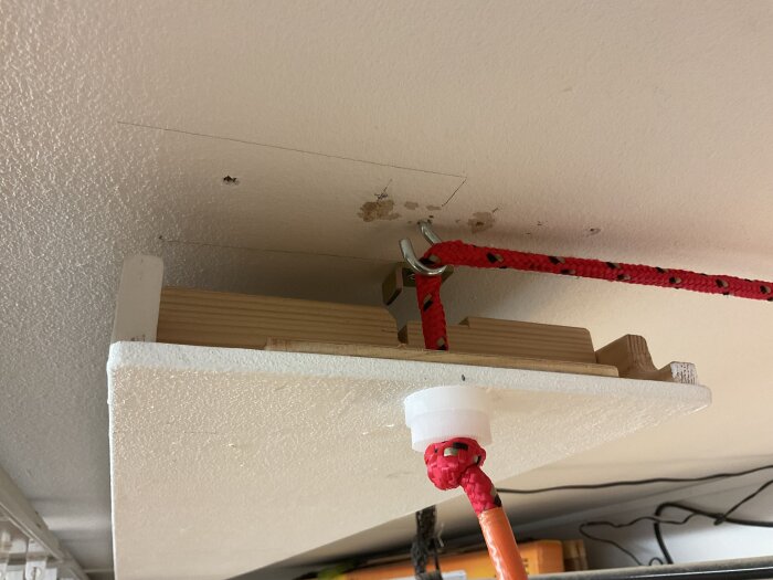 Fästanordning för verktygslåda med röda rep, under tak, med simpel hemmagjord konstruktion.