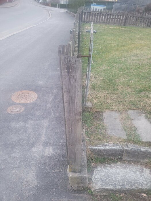 Gamla trästaketet längs med sluttande betongsockel bredvid en gräsmatta och en väg.