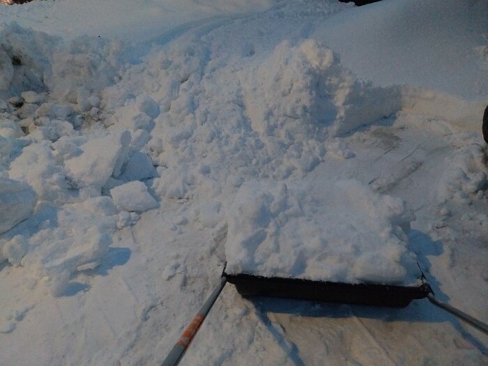 Snöskottning utanför hus med en stor hög av bortschaktad snö och en snöskyffel i förgrunden.