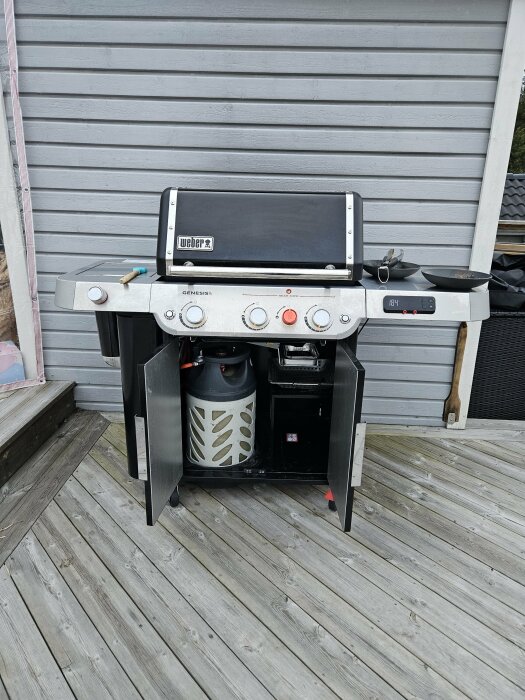 Weber gasolgrill med stängt lock på träaltan, grillverktyg och gasoltub synlig.