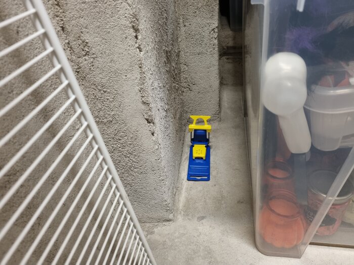 En liten blå och gul verktygsvagn mellan en vit hylla och en transparent plastlåda mot en betongvägg.