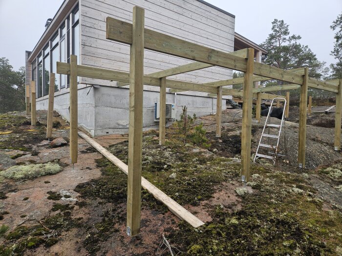 Stolpar och bärregler av trä monterade för grundläggning av en altan, nära en byggnad på klippig mark.