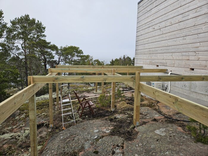 Trästomme av stolpar och balkar under uppbyggnad för en altan, bredvid en träbeklädd byggnad, med en stege och naturen i bakgrunden.