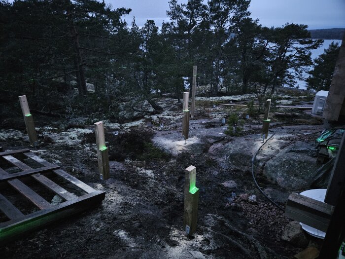 Byggprojekt i skymning med stolpar och laserpass linjer på en klippig terräng nära vatten.