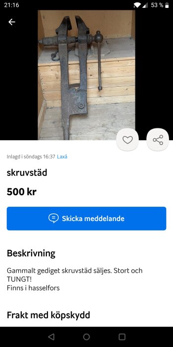 Gammal stor skruvstäd i metall på en träbänk, till försäljning för 500 kr, text anger "TUNGT! Finns i hasselfors".