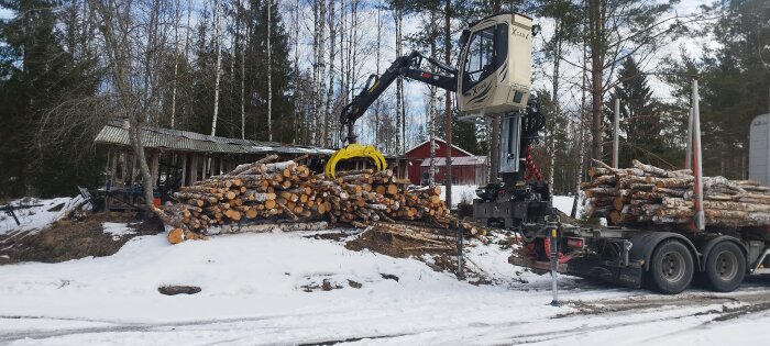 Gripklo på skogsmaskin lyfter björkved från lastbil vintertid med snö och skog i bakgrunden.