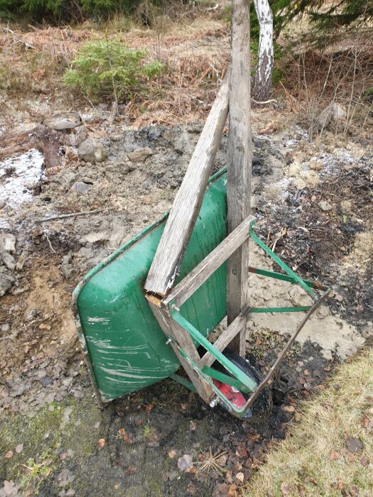 En trasig grön skottkärra med avbrutet handtag vält i lerigt och grusigt område vid en grävd trappa.