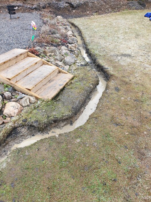 Grävd ränna i trädgård framför trätrapp med vattenpölar och lerig mark, omgiven av gräs och stenar.