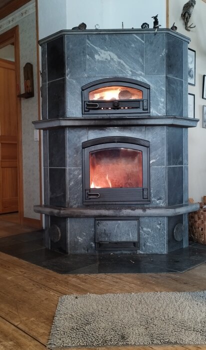 Tulikivi-kakelugn av sten med eld och glöd synlig i eldstaden, placerad i ett hörn av ett kök.
