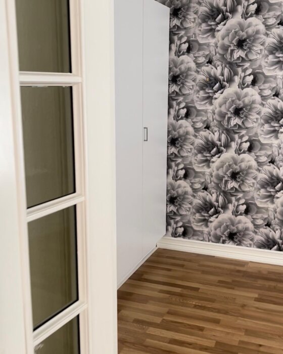 Vägg med grått blommig tapet, vitmålad del och trägolv syns i ett hörn av rummet.