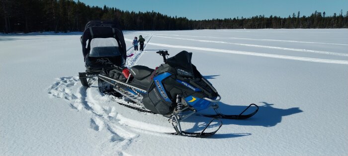 Snöskoter parkerad på en snötäckt sjö med två personer och en pulka i bakgrunden.
