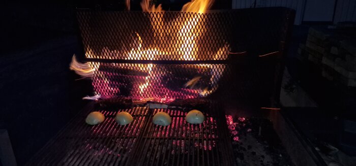 Vedeldad grill med flammor bakom ett metallgaller och fyra halverade lökar på grillgallret i kvällsbelysning.