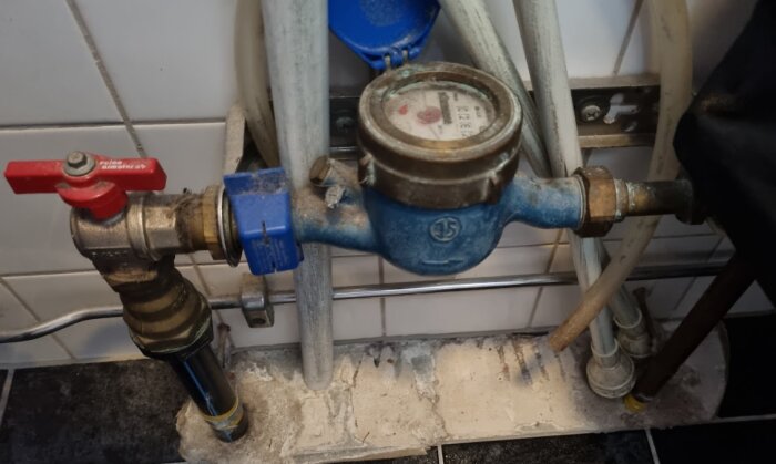 Gammal vattenmätare installerad genom golvet med synliga rör och stängningsventiler i ett badrum.