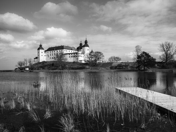 Svartvit bild av Läckö slott vid vattenkant med brygga och vass i förgrunden.