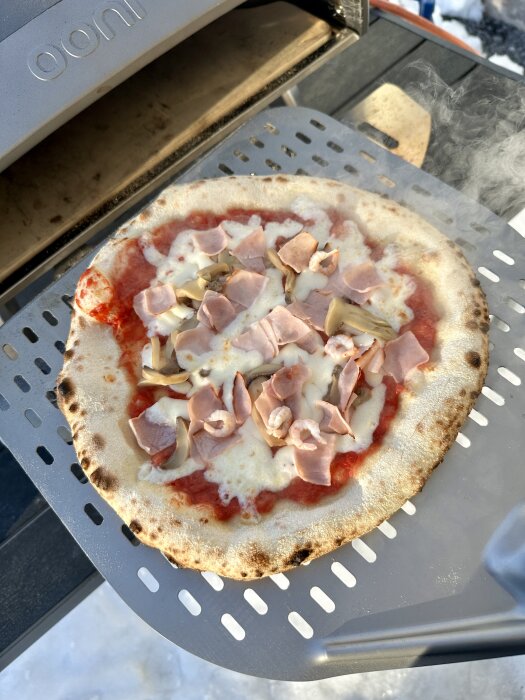 Nybakad pizza med skinka och champinjoner på en spatel framför en portabel pizzaugn.