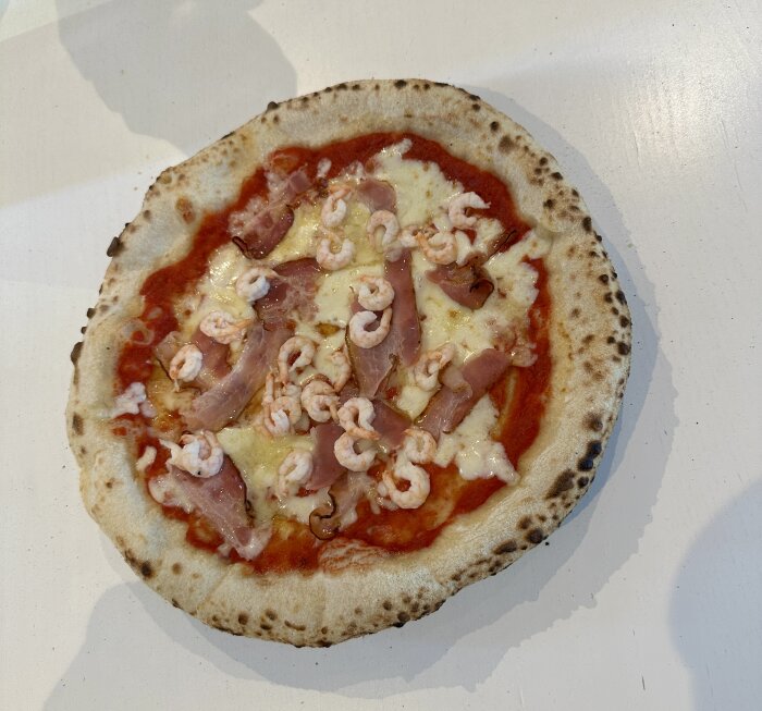 Hemgjord pizza med tomatsås, ost, skinka och räkor på en ljus bakgrund.