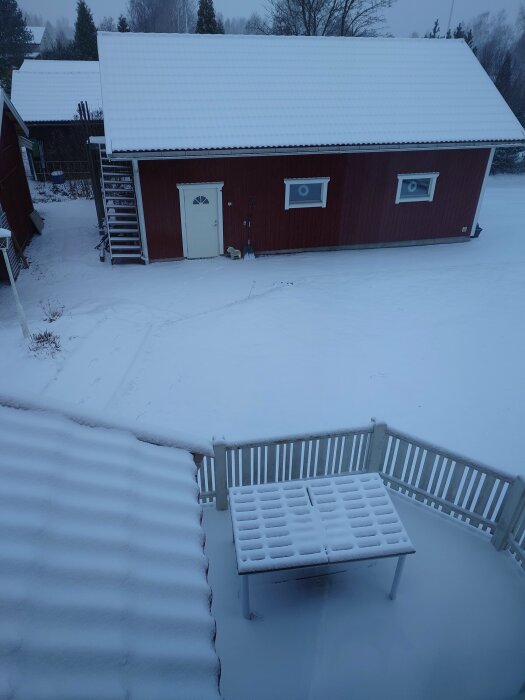 Röd träbyggnad med vit dörr och snötäcke på taket, omgiven av snöklädd mark och en trädgårdsmöbel täckt av snö.