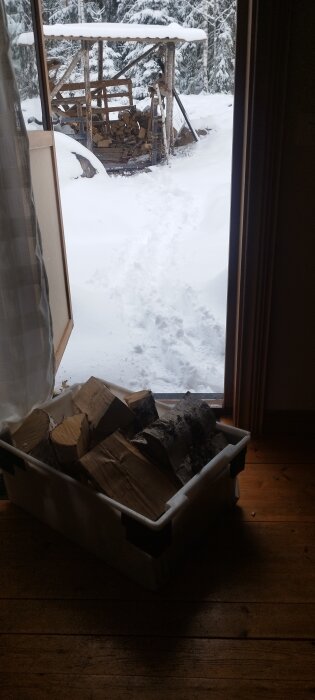 En låda med vedblock inne och en vedbod täckt med snö sedd genom en öppen dörr.