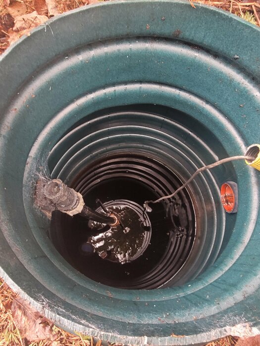Öppning av en brunnskammare med en kabel som hänger ner i vatten, omgiven av en grön plastkant.