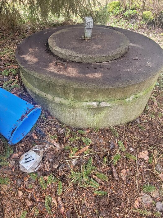 Betonbrunn med lock i skogen, blå hink och en kabel inslagen i plastpåsar på marken.