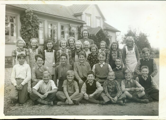 Grupp av leende barn framför gammalt hus, svartvit bild från mitten av 1900-talet.