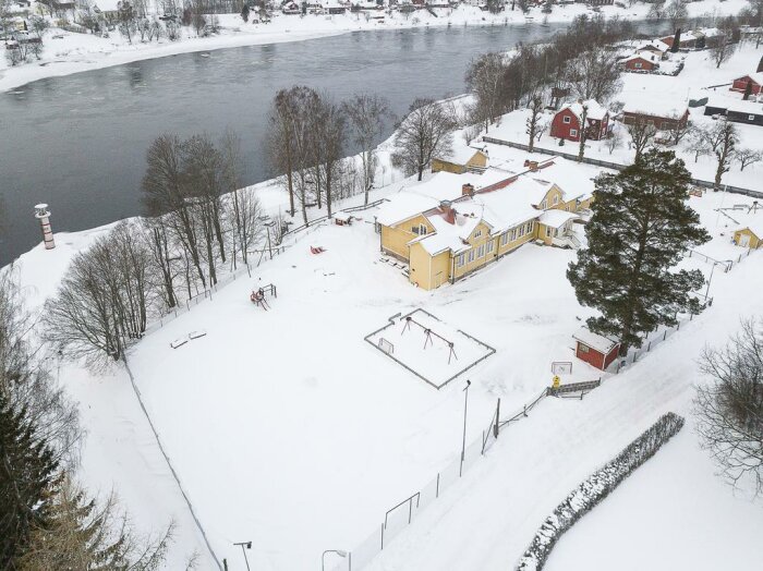 Flygbild över gult hus med utbyggnader och tillhörande småbyggnader omgivet av snö och stängsel nära en flod.