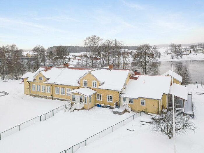 Gult hus med utbyggnader och verandor omgivet av snö och stängsel, nära en frusen flod.
