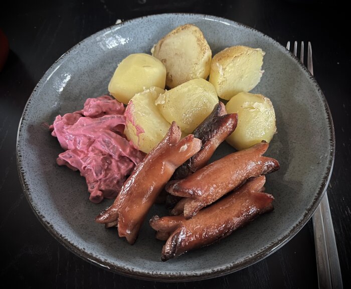 En tallrik med prinskorv, kokt potatis och rödbetssallad på ett mörkt bord, redo för måltid.