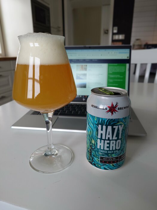 Ölglas fyllt med hazy öl och en burk märkt "Hazy Hero" framför en laptop på ett bord.
