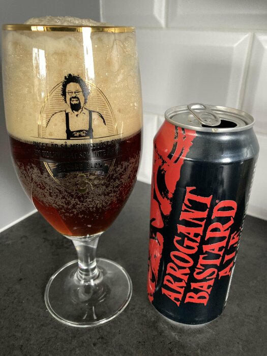 Ett glas öl med tjockt skum bredvid en burk med texten "Arrogant Bastard Ale" på en köksbänk.