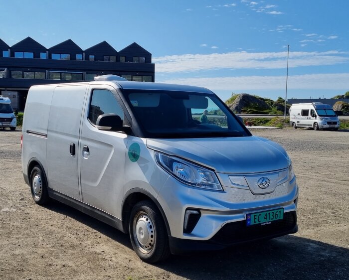 En eldriven Maxus skåpbil parkerad utomhus med andra fordon och byggnader i bakgrunden.