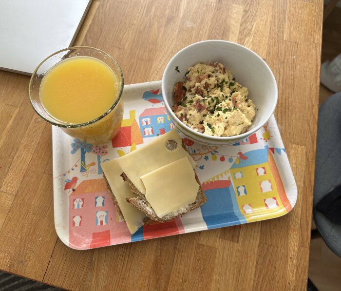 Frukostbricka med apelsinjuice, äggröra med sidfläsk och persilja, ostsmörgås på ett färgglatt papper.